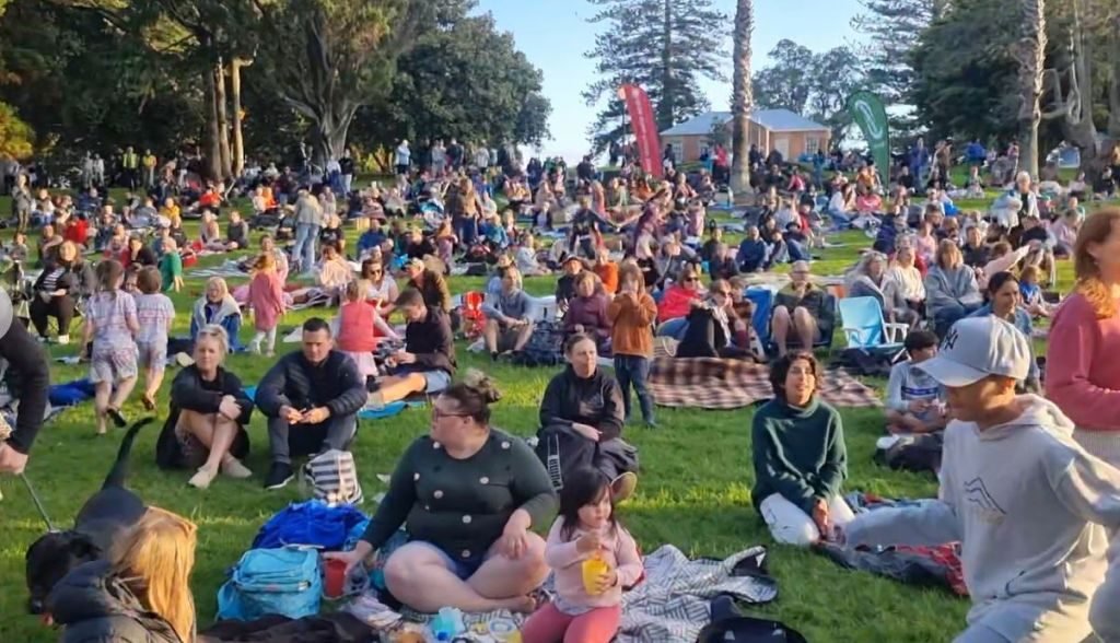 A big crowd sitting on the grass at an evening public event