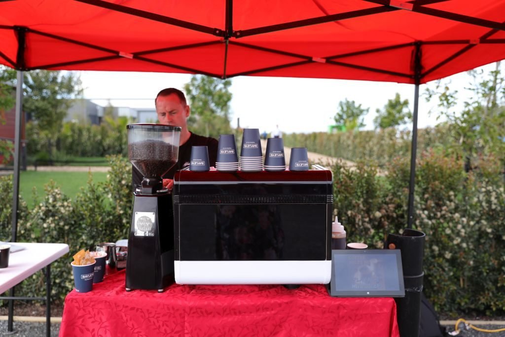 Espresso machine and grinder set up under a red gazebo with the barista behind